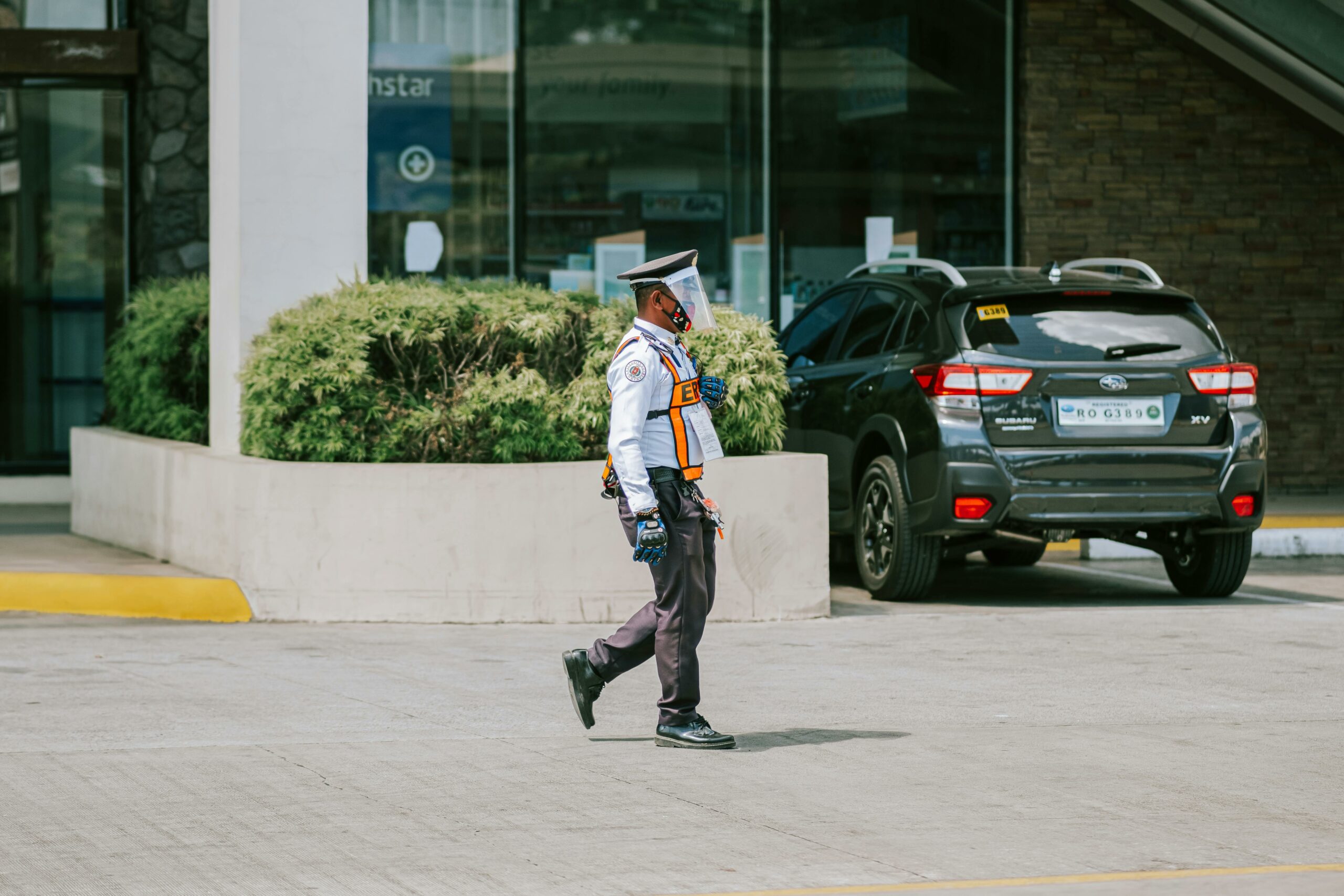A security guard wearing a mask and gloves walks by a parked car outdoors, ensuring safety.